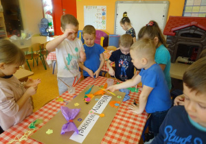 Dzieci dekorują plakat, naklejają kolorowe kwiaty z papieru i bibuły.